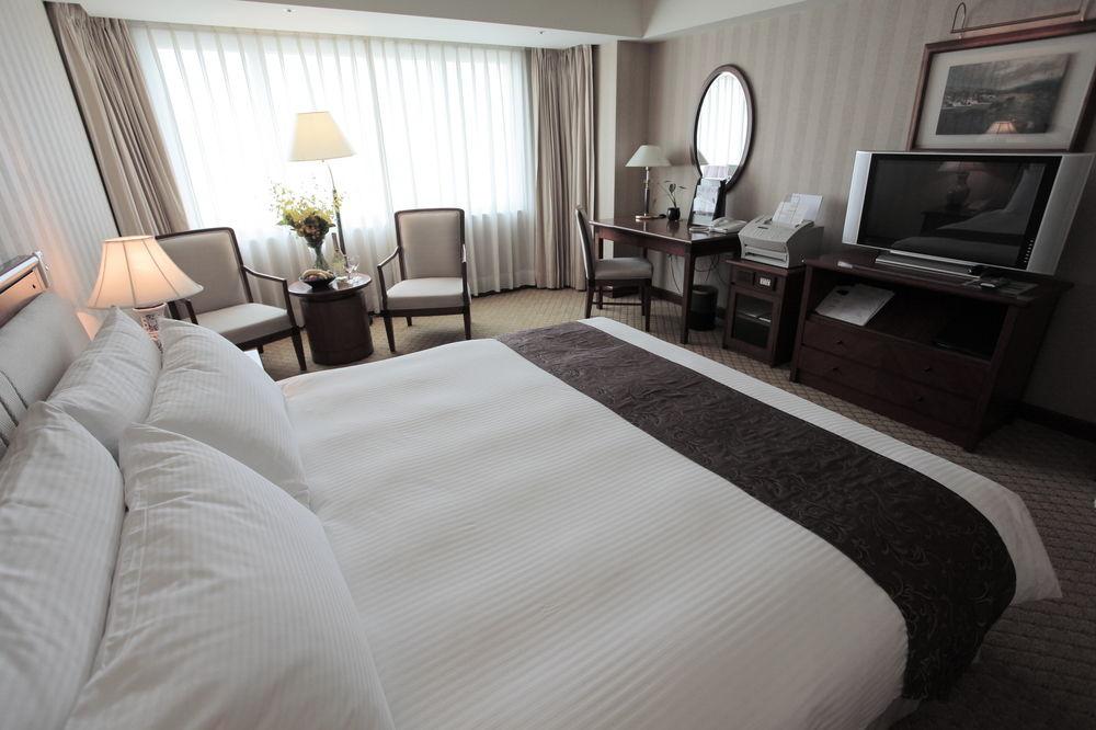 Evergreen Laurel Hotel - Taichung Luaran gambar The photo shows a well-furnished hotel room. In the foreground, there is a large bed with white bedding and a dark-colored throw at the foot. Beside the bed, there is a bedside lamp. 

In the background, natural light filters through large windows co