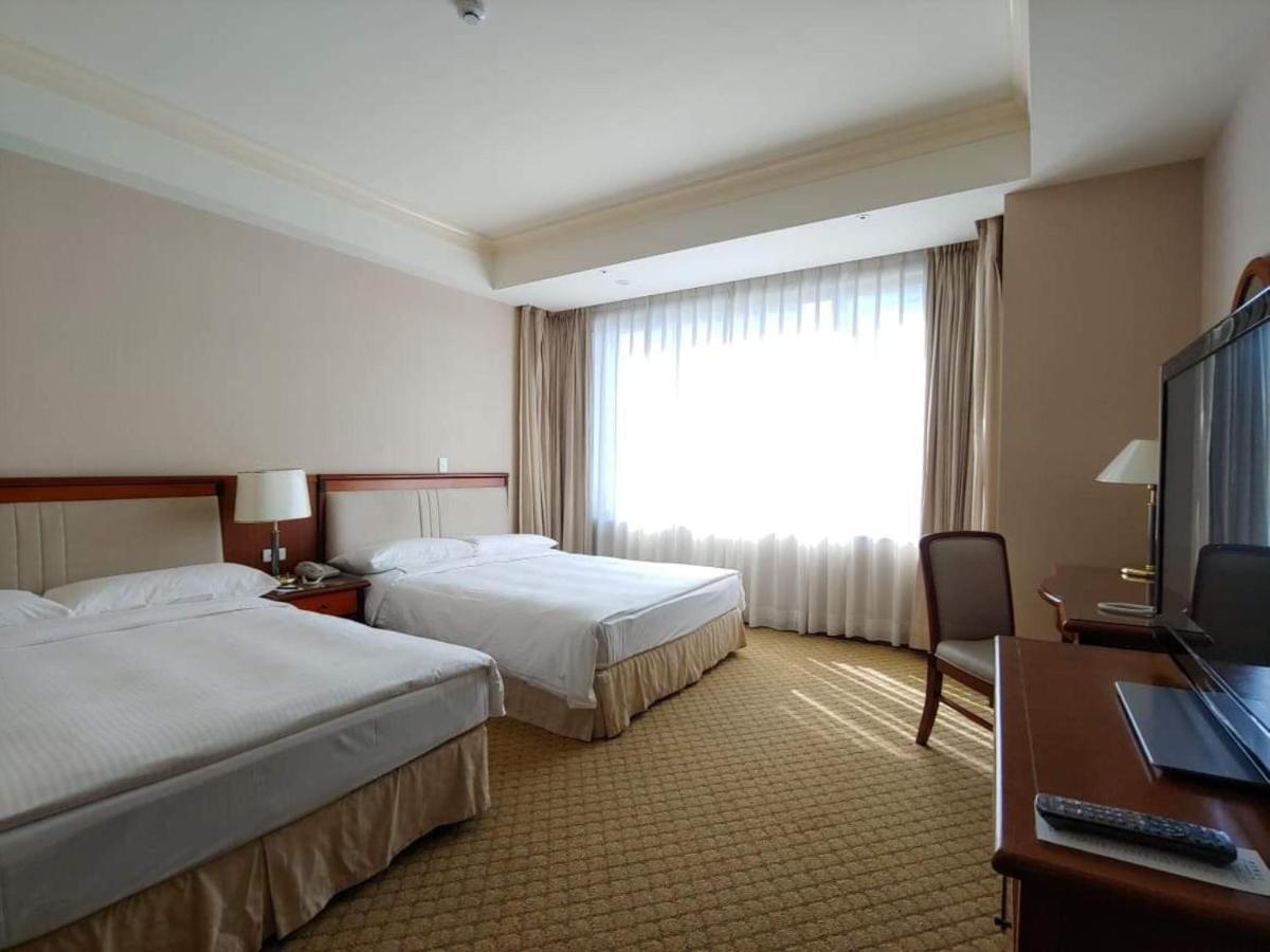 Evergreen Laurel Hotel - Taichung Luaran gambar The image shows a hotel room featuring two single beds with white bedding. There is a large window with sheer curtains letting in natural light, and the walls are painted in neutral tones. A wooden desk with a chair is positioned near the window, and