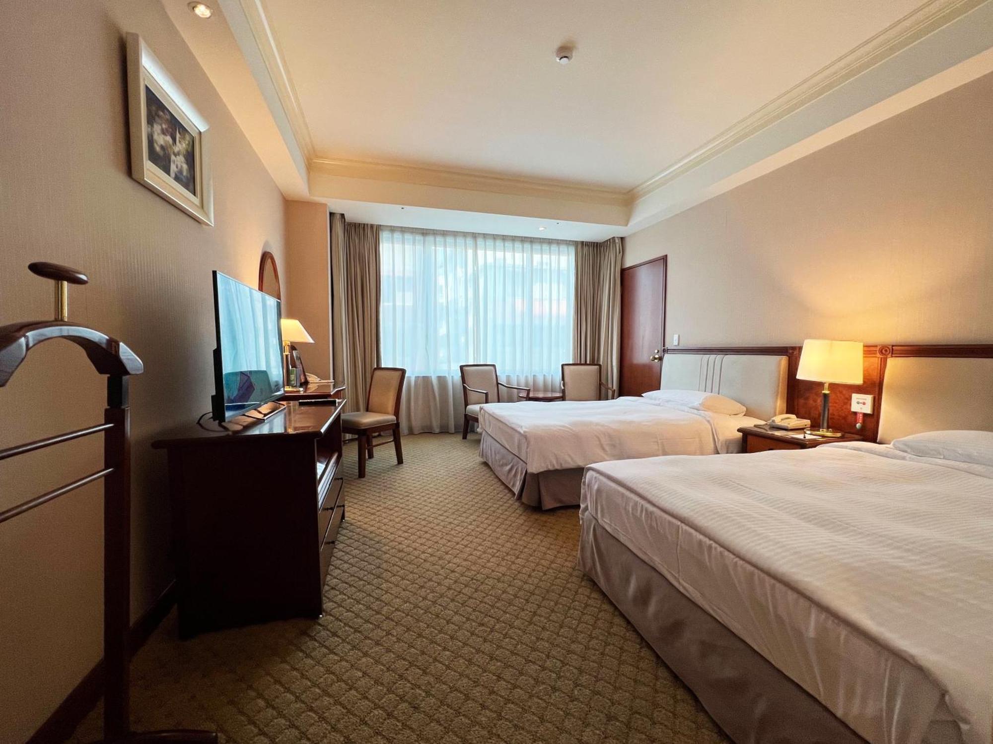 Evergreen Laurel Hotel - Taichung Luaran gambar The photo shows a hotel room featuring two twin beds placed parallel to each other. The room has a warm color palette and includes a large window with sheer curtains allowing natural light to illuminate the space. There is a wooden desk and two chair