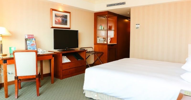 Evergreen Laurel Hotel - Taichung Luaran gambar The photo shows a hotel room featuring a neatly made bed with a white blanket. On one side, there is a wooden desk with a chair, and a flat-screen television is mounted nearby. An accent cabinet is visible, displaying various items. The walls are pai