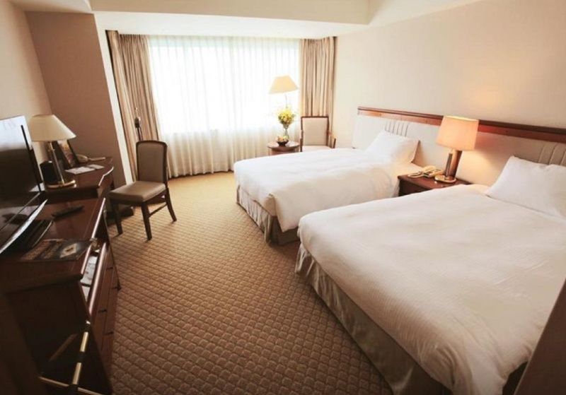 Evergreen Laurel Hotel - Taichung Luaran gambar The photo shows a hotel room featuring two twin beds with white bedding. There's a chair positioned near one of the beds and a desk with a computer setup against one wall. Large windows allow natural light to enter the room, and light-colored curtain