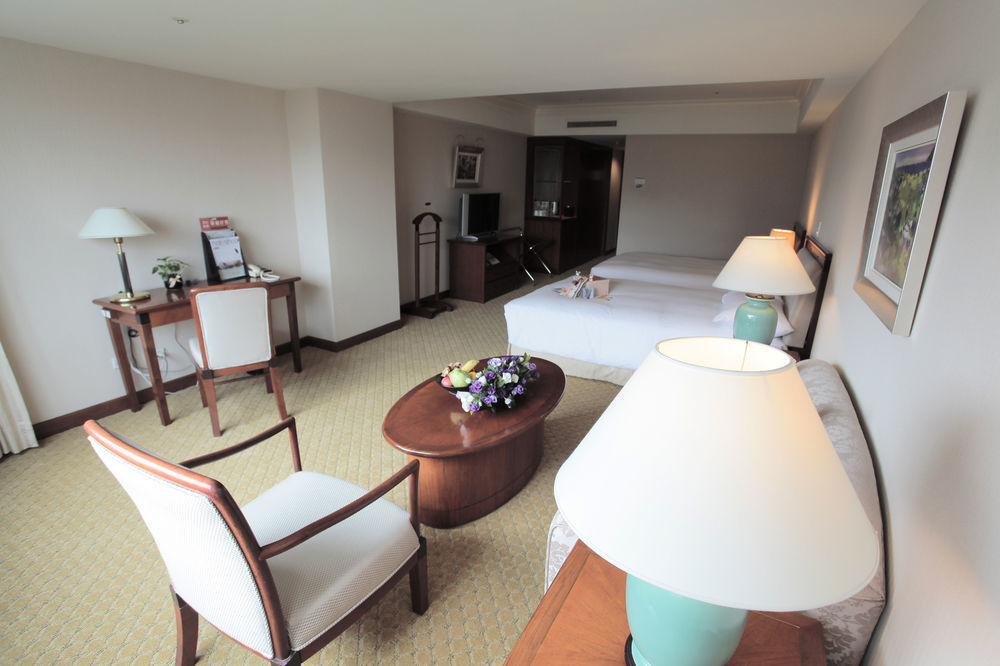 Evergreen Laurel Hotel - Taichung Luaran gambar The photo shows an interior view of a hotel room. It features two beds, a coffee table with a small decorative arrangement of flowers, and a comfortable chair. There's a desk and a lamp, along with another larger lamp on the table. In the background,