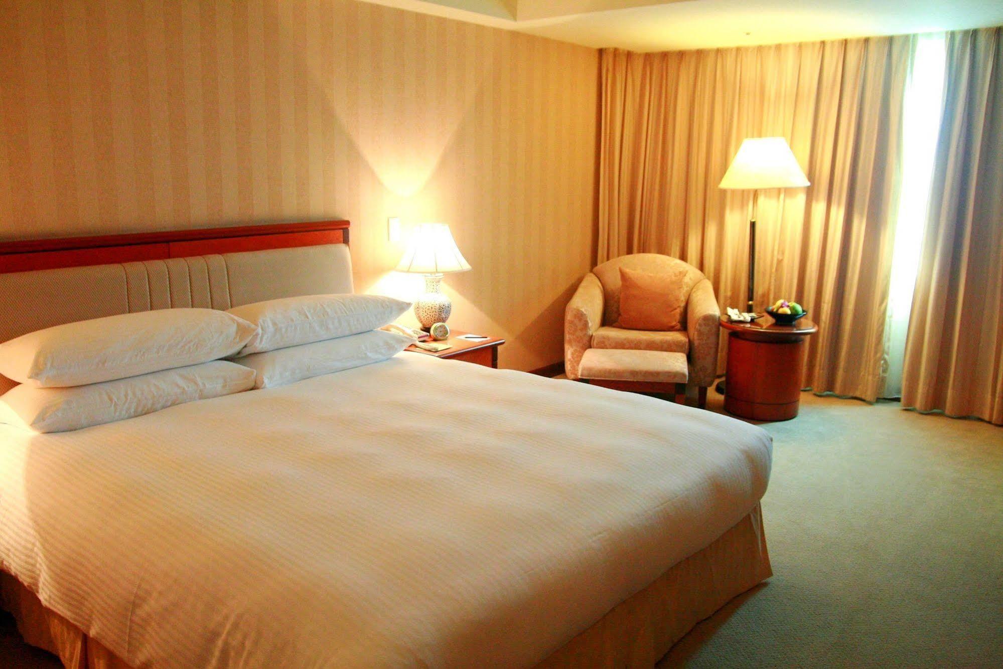 Evergreen Laurel Hotel - Taichung Luaran gambar The photo shows a hotel room containing a large bed with white linens, flanked by two bedside lamps. To one side, there is a cozy armchair and a small side table with a fruit bowl or snacks. The walls are decorated in a soft, neutral color, and there