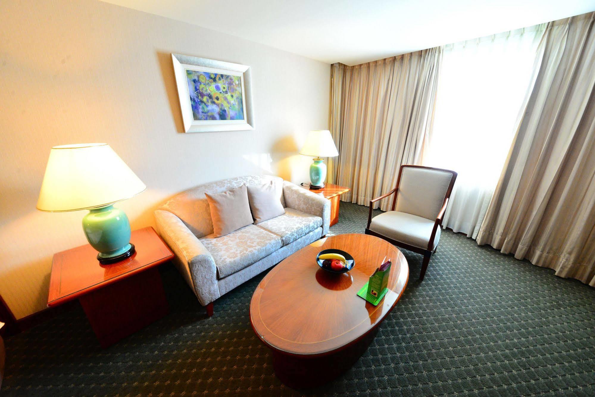 Evergreen Laurel Hotel - Taichung Luaran gambar The photo shows a cozy living space, likely a hotel room or a lounge area. It features a light-colored sofa with cushions, a wooden coffee table in the center, and a comfortable armchair beside it. There are two lamps with turquoise bases on side tab
