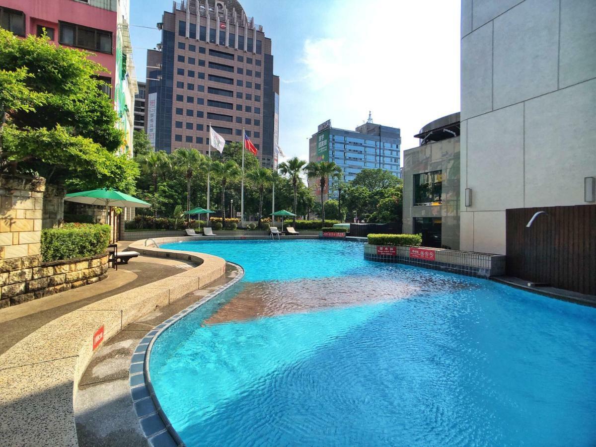 Evergreen Laurel Hotel - Taichung Luaran gambar The photo shows a swimming pool set in a modern urban environment. The pool has a curved edge and is surrounded by greenery, including trees and umbrellas for shade. In the background, there are several tall buildings, indicating a cityscape, with so