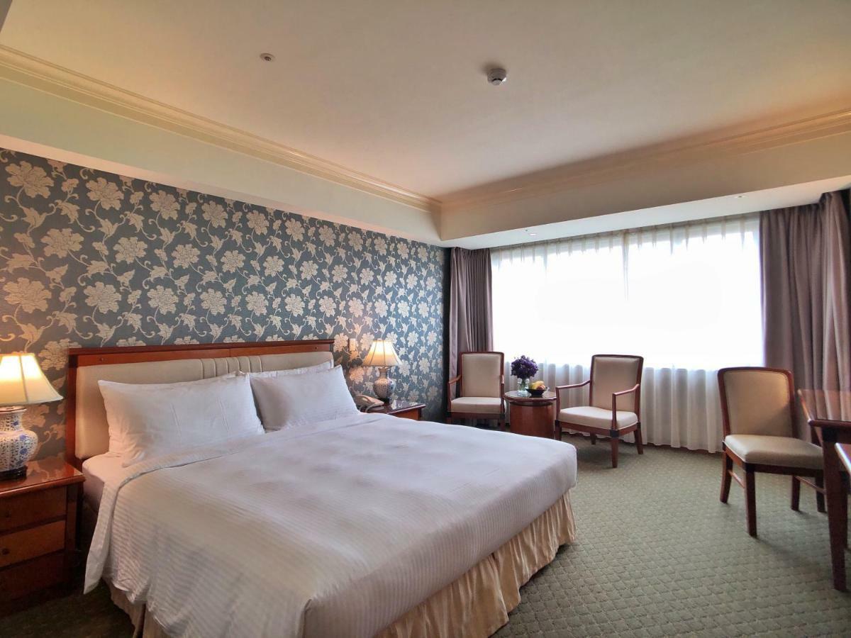 Evergreen Laurel Hotel - Taichung Luaran gambar The photo shows a hotel room that features a large bed with a white bedspread positioned against a decorative floral wallpaper. There are two bedside lamps on wooden nightstands flanking the bed. To the side of the room, there are two chairs and a sm