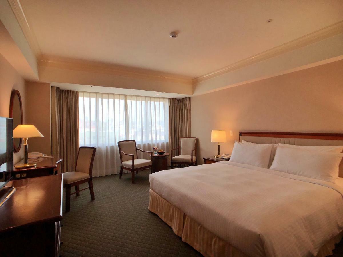 Evergreen Laurel Hotel - Taichung Luaran gambar The photo shows a hotel room featuring a large bed with white linens, a wooden bedside table with a lamp, and a flat-screen television. There are two chairs with a small table next to them positioned by a large window, which has sheer curtains lettin