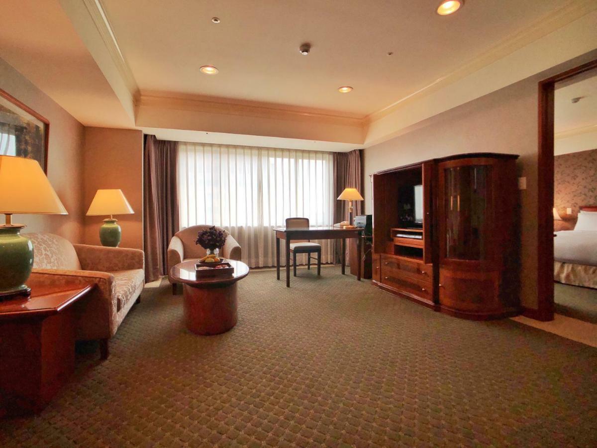 Evergreen Laurel Hotel - Taichung Luaran gambar The photo shows a cozy hotel suite or living area. There is a sofa and an armchair arranged around a small coffee table. A desk with a chair is positioned near a large window covered with sheer curtains that allow natural light to enter the room. Two
