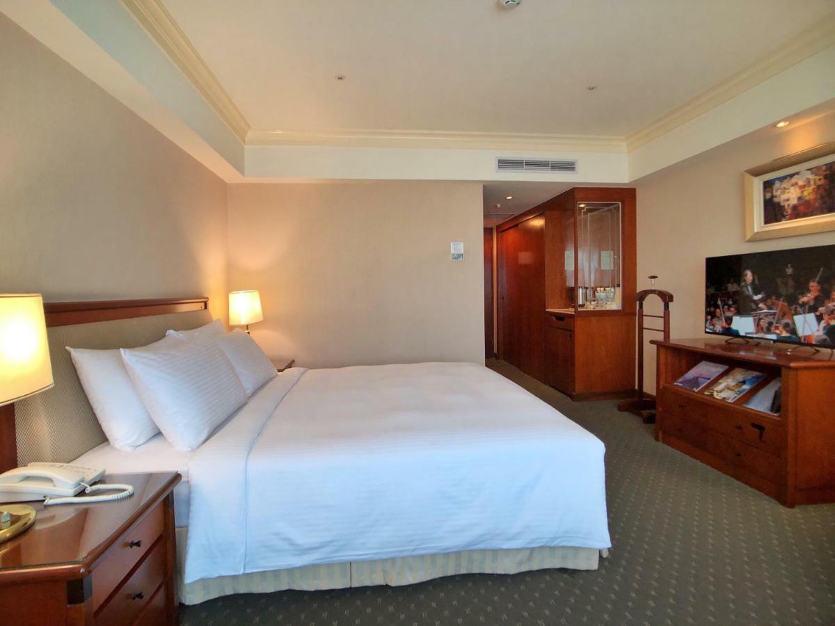 Evergreen Laurel Hotel - Taichung Luaran gambar The photo shows a hotel room featuring a comfortable bed with white linens and pillows, positioned against a light-colored wall. There is a wooden bedside table with a lamp and an open book next to the bed. On the opposite side, there’s a large flat-