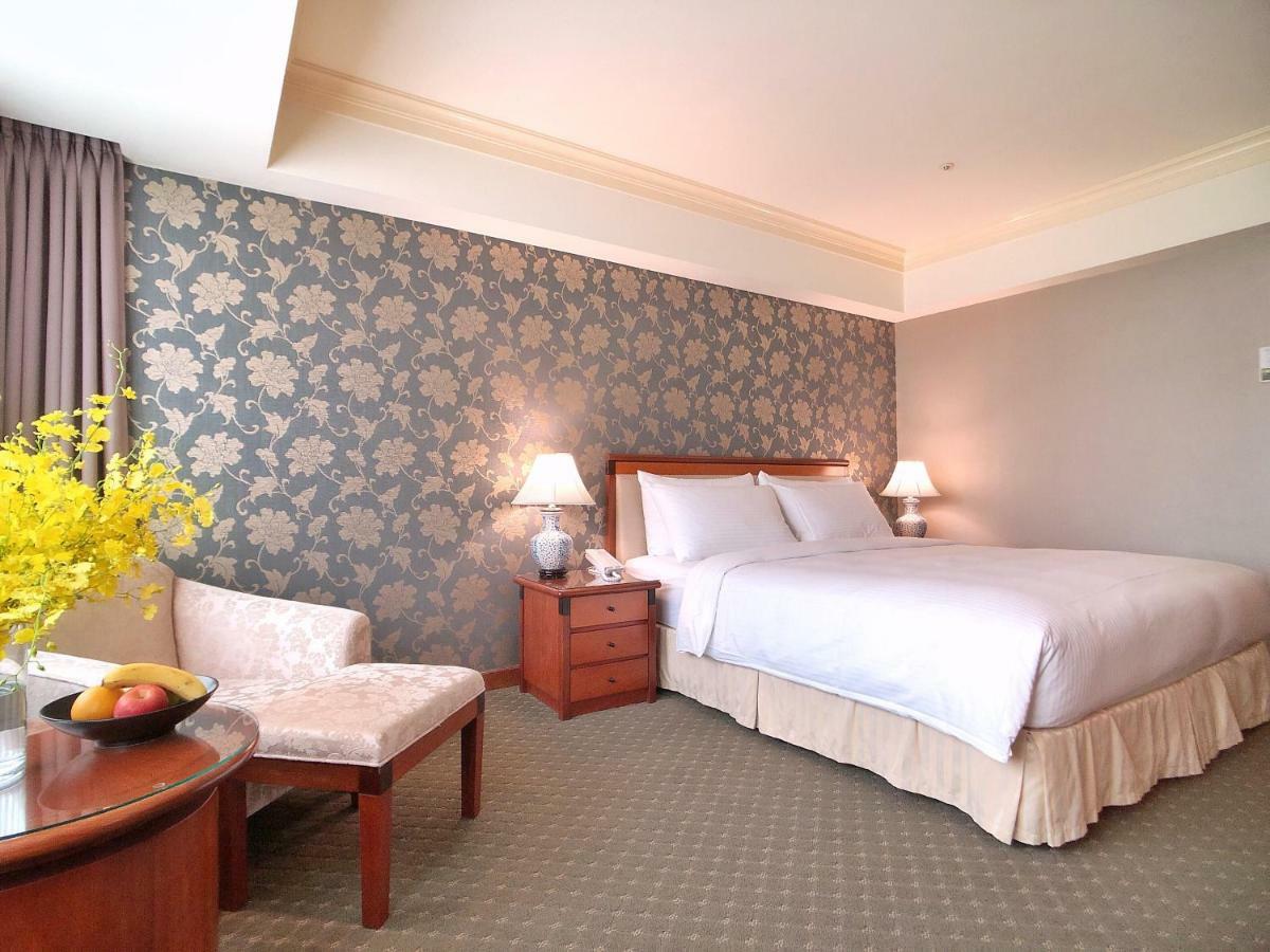 Evergreen Laurel Hotel - Taichung Luaran gambar The photo shows a well-decorated hotel room. It features a large bed with white bedding, situated against a patterned wall with floral designs. On either side of the bed are bedside tables with lamps. There is a small seating area that includes a cha