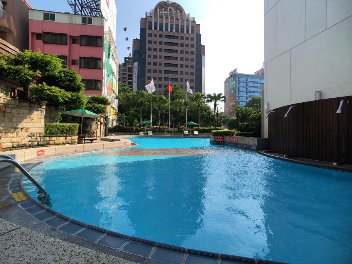 Evergreen Laurel Hotel - Taichung Luaran gambar The photo shows a swimming pool area set in an outdoor environment. The pool has a clear blue water and a gently curving shape. Surrounding the pool are some green trees and plants, along with a few umbrellas providing shade. In the background, there