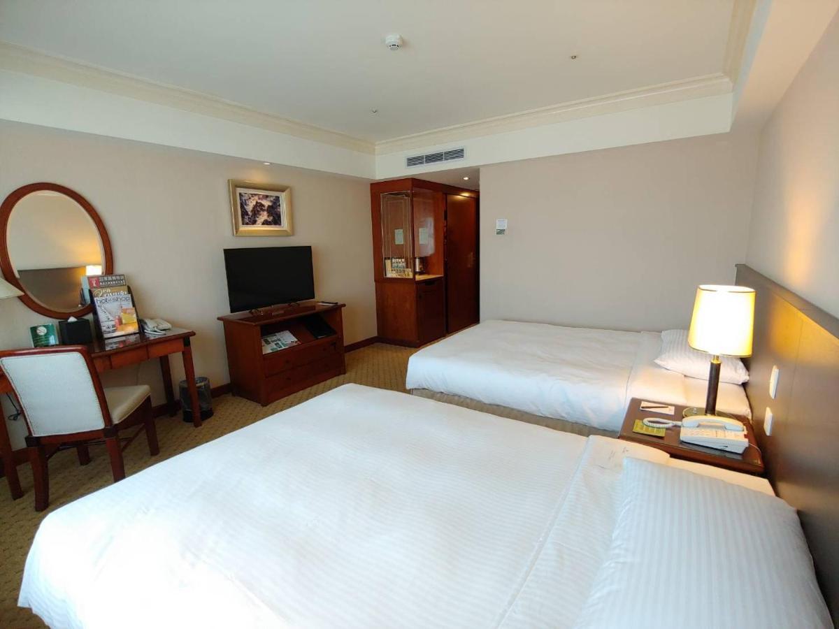 Evergreen Laurel Hotel - Taichung Luaran gambar The photo shows a hotel room with two beds, neatly made with white linens. There is a desk and chair in one corner, accompanied by a flat-screen TV mounted on the wall. A large mirror and a wooden cabinet are also visible. The room is illuminated by 